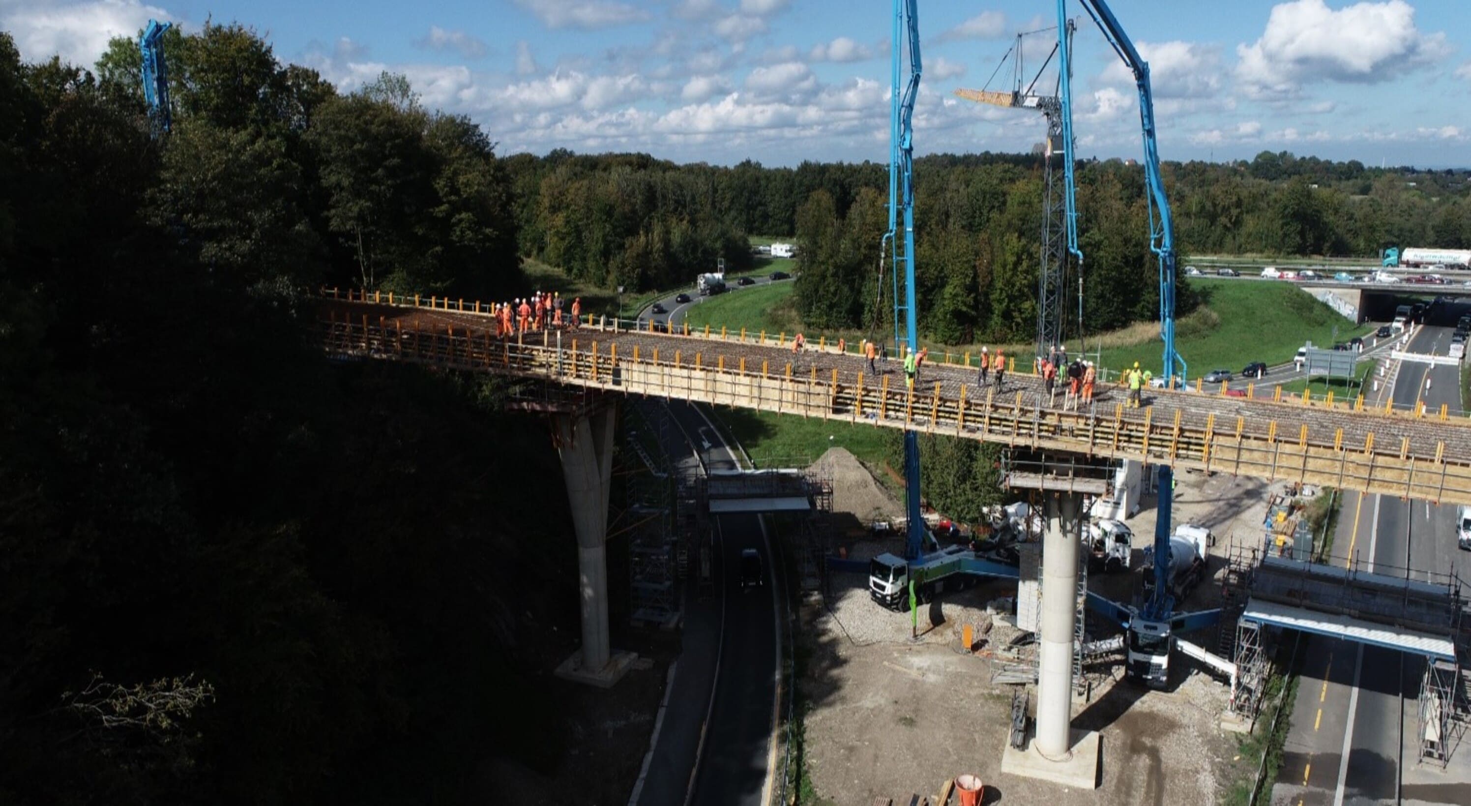 Eine Brücke im Bau wird aus der Vogelperspektive gezeigt. Die Brückenkonstruktion verläuft über eine Straße und wird von mehreren Betonpfeilern getragen. Bauarbeiter in orangefarbener Schutzkleidung sowie eine große blaue Betonpumpe sind auf der Brücke im Einsatz. Im Hintergrund sind Straßen, Autos und viel Grün zu erkennen.

                                      